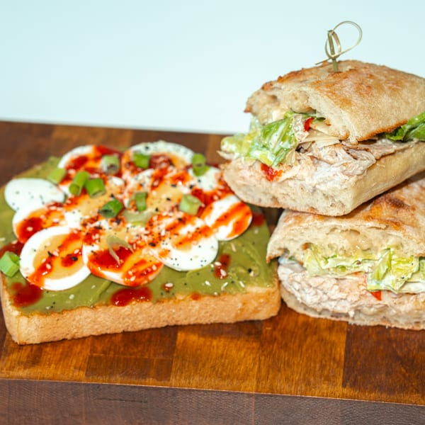 Avocado toast with sliced eggs and two chicken salad sandwiches on a wooden board.