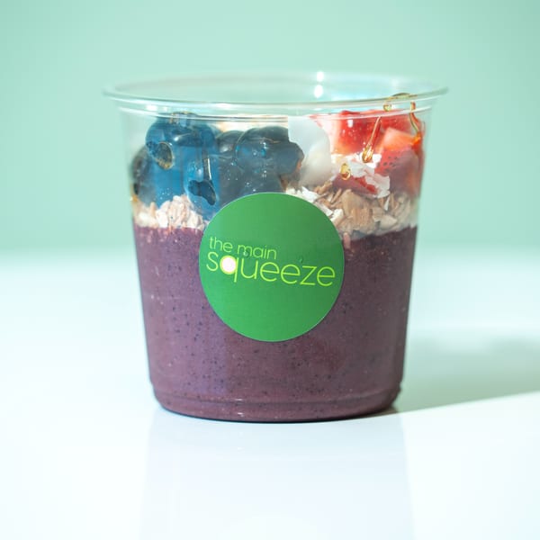 A Main Squeeze smoothie cup with purple smoothie, blueberries, strawberries, granola, and coconut flakes.